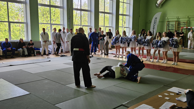 Sporta zālē uz tatami cīnās divi vīrieši kimono, notiek džudo cīņa. Blakus notiekošo vēro tiesnesis, gar tatami malu, gaidot savu kārtu citi sacensību dalībnieki. Blakus tatami paklājiem rindā stāv karsējmeitenes ar zili-baltiem pušķiem rokās