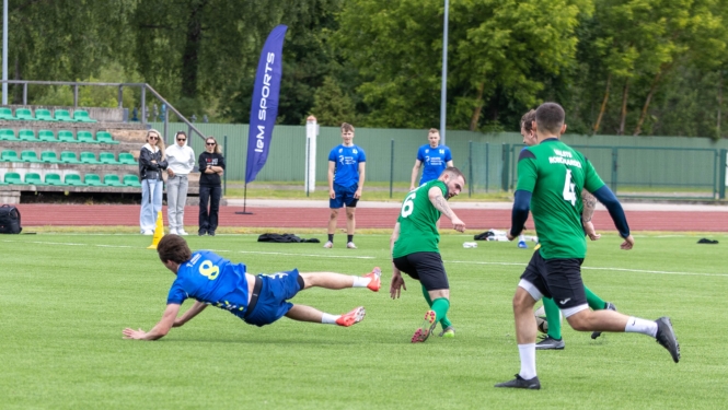 Futbola laukumā vīrieši spēlē futbolu. Laukuma malā trīs sievietes vēro spēli.