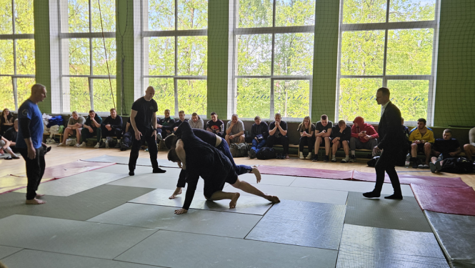 Sporta zālē uz tatami cīnās divi vīrieši, notiek džudo cīņa. Blakus notiekošo vēro tiesnesis un citi sacensību dalībnieki.