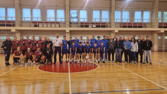 Sporta spēļu zālē kopbildei sastājušās trīs telpu futbola sacensību uzvarētāju komandas – pirmās un otrās vietas ieguvēji ģērbti sporta spēļu formās. Komandu dalībniekiem un treneriem kaklā medaļas, rokās vai priekšā uz grīdas – komandas izcīnītais kauss.