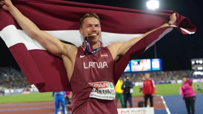 sportists stadionā ar Latvijas karogu abās paceltās rokās medaļa mutē