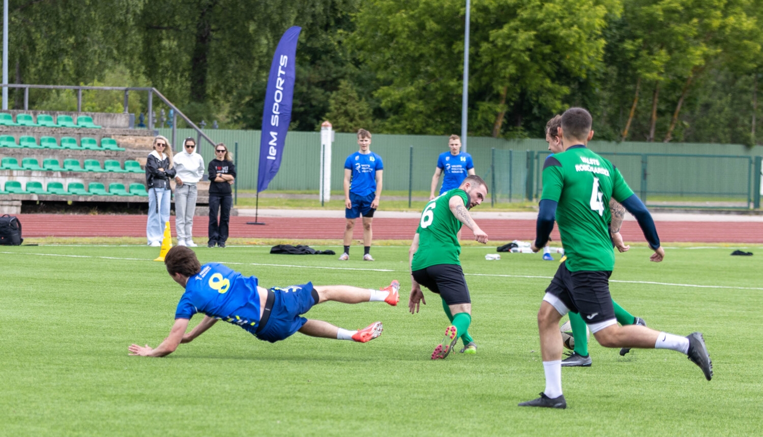 Futbola laukumā vīrieši spēlē futbolu. Laukuma malā trīs sievietes vēro spēli.