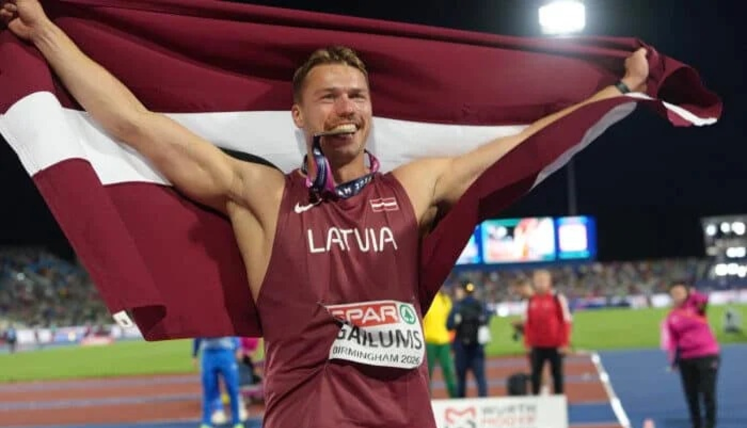 sportists stadionā ar Latvijas karogu abās paceltās rokās medaļa mutē