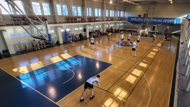 Sporta spēļu zāle centrā novilkts volejbola tīkls Valsts policijas apvienotā komanda dzeltenos kreklos un melnos šortos un Valsts policijas koledžas komanda pelēkos kreklos ar melniem šortiem spēlē volejbolu koledžas spēlētājs gatavojas padot servi