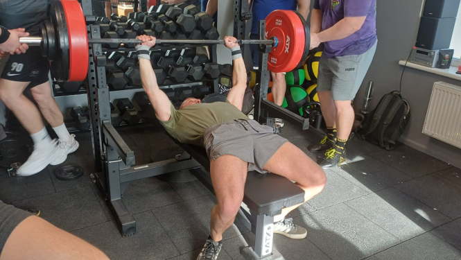 Sacensību dalībnieks spiež svara stieni guļus uz muguras, divi citi dalībnieki drošina, turot stieni aiz galiem