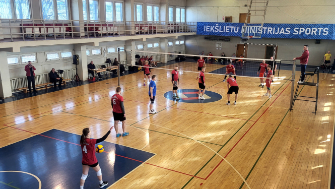 No augšas uzņemta fotogrāfija atklāj intensīvu volejbola maču plašā, saules apgaismotā sporta zālē, kas met asas ēnas uz koka grīdas. Abās volejbola tīkla pusēs pa sešiem spēlētājiem: pieci vīrieši nogaidošās pozās gaida kad sieviete servēs bumbu. Laukuma centrā, uz paaugstinātas platformas tieši pie tīkla, stāv tiesnesis ar svilpi mutē, uzmanīgi seko katram spēles momentam. 