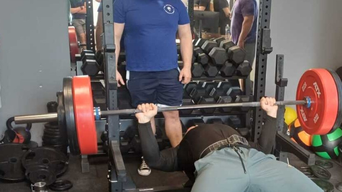 Sporta zālē veic guļus spiešanu ar stieni uz sola.Fonā hanteles.