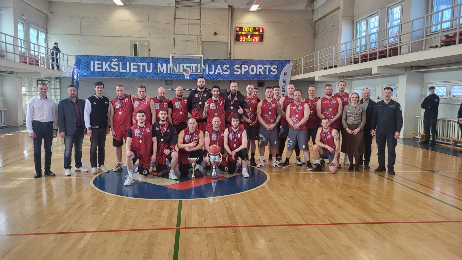 Sporta zālē kopbildē divas no laureātu komandām – pirmās vietas ieguvēji Valsts policija Rīga un VUGD komanda. Spēlētājiem kaklā zelta un sudraba medaļas, uz grīdas priekšā stāv ceļojošais čempionu kauss un pirmās vietas ieguvēju kauss.