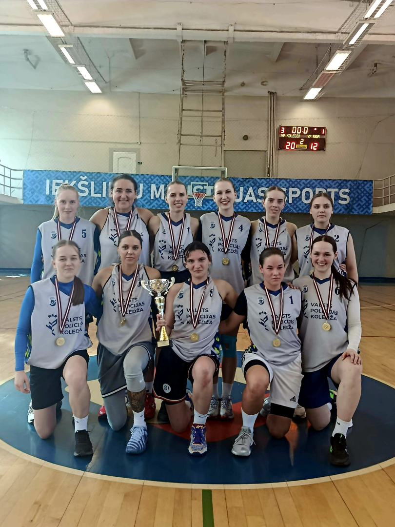 Basketbola sacensību čempiones – Valsts policijas koledžas komanda sporta zālē, visām spēlētājām kaklā zelta medaļas, vienai spēlētājai – komandas izcīnītais kauss.