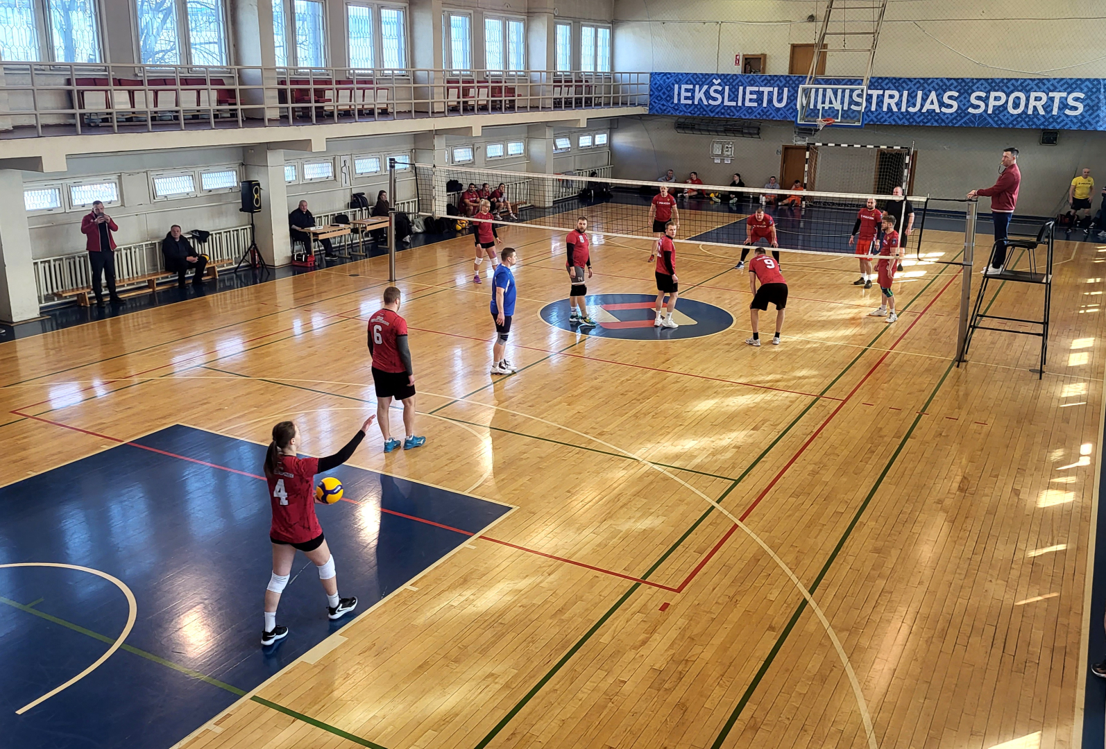 Sporta zāle centrā novilkts volejbola tīkls uz paaugstinājuma blakus stāv tiesnesis fonā redzams tumši zils baneris ar uzrakstu Iekšlietu ministrijas sports 2 komandas sarkanos kreklos un šortos spēlē volejbolu spēlētāja gatavojas veikt servi