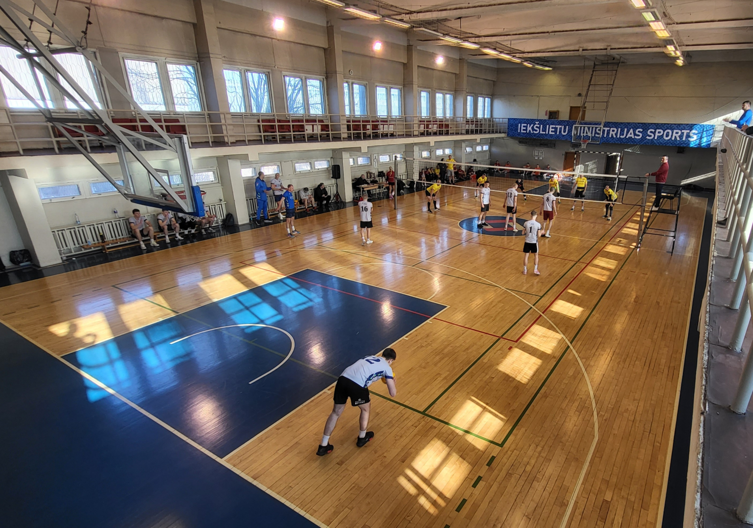 Sporta spēļu zāle centrā novilkts volejbola tīkls Valsts policijas apvienotā komanda dzeltenos kreklos un melnos šortos un Valsts policijas koledžas komanda pelēkos kreklos ar melniem šortiem spēlē volejbolu koledžas spēlētājs gatavojas padot servi