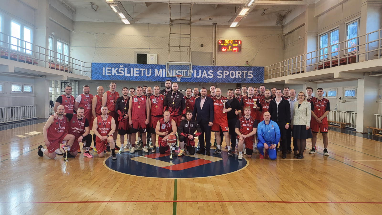 Valsts policijas čempionāta sacensību basketbolā laureātu un apbalvotāju kopbilde sporta zālē. Spēlētāji sarkanās formās ar medaļām un kausiem stāv un tup uz laukuma, fonā redzams basketbola grozs un rezultātu tablo.