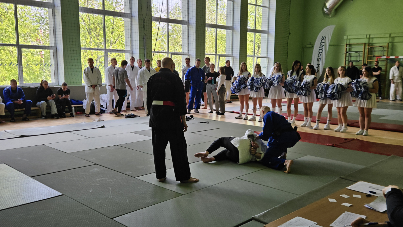 Sporta zālē uz tatami cīnās divi vīrieši kimono, notiek džudo cīņa. Blakus notiekošo vēro tiesnesis, gar tatami malu, gaidot savu kārtu citi sacensību dalībnieki. Blakus tatami paklājiem rindā stāv karsējmeitenes ar zili-baltiem pušķiem rokās