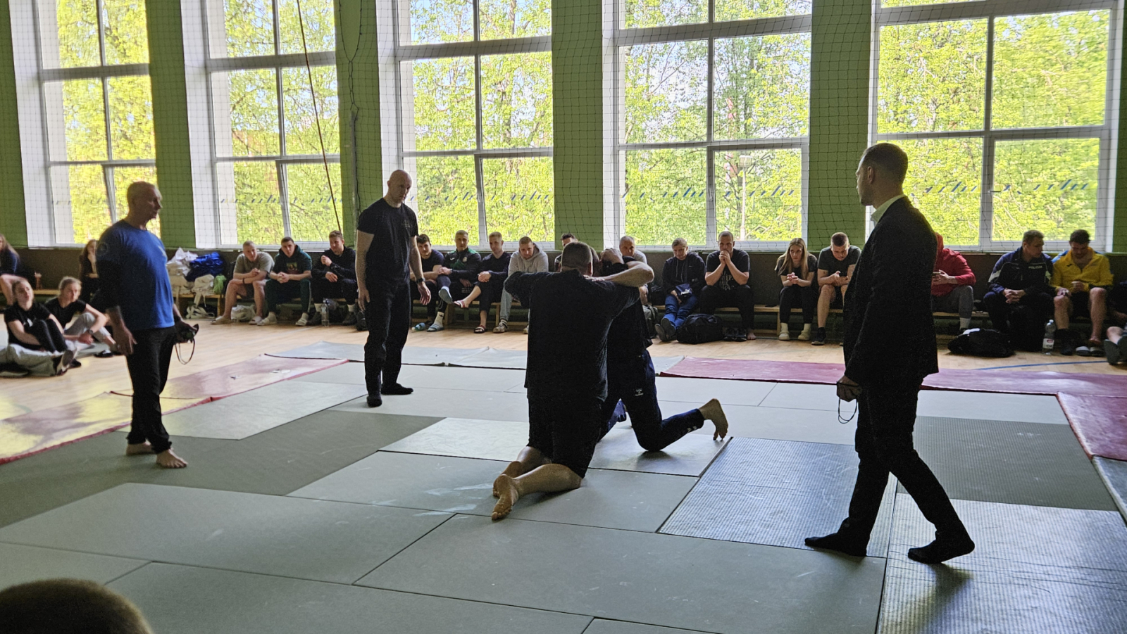 Sporta zālē uz tatami uz ceļiem cīnās divi vīrieši, notiek džudo cīņa. Blakus notiekošo vēro tiesnesis un citi sacensību dalībnieki.