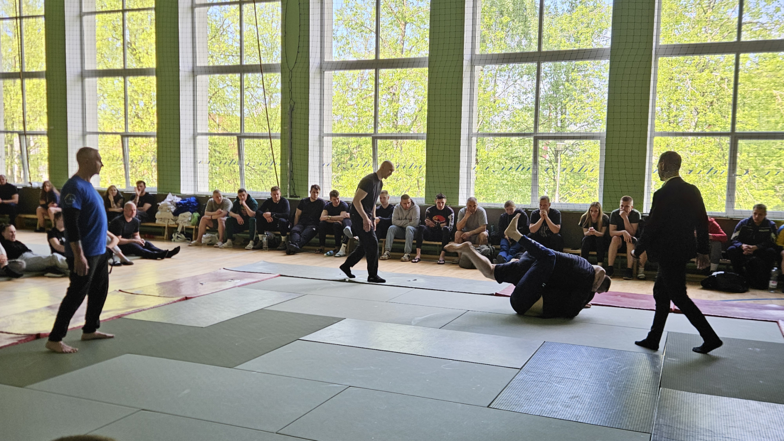 Sporta zālē uz tatami cīnās divi vīrieši, notiek džudo cīņa. Blakus notiekošo vēro tiesnesis un citi sacensību dalībnieki.