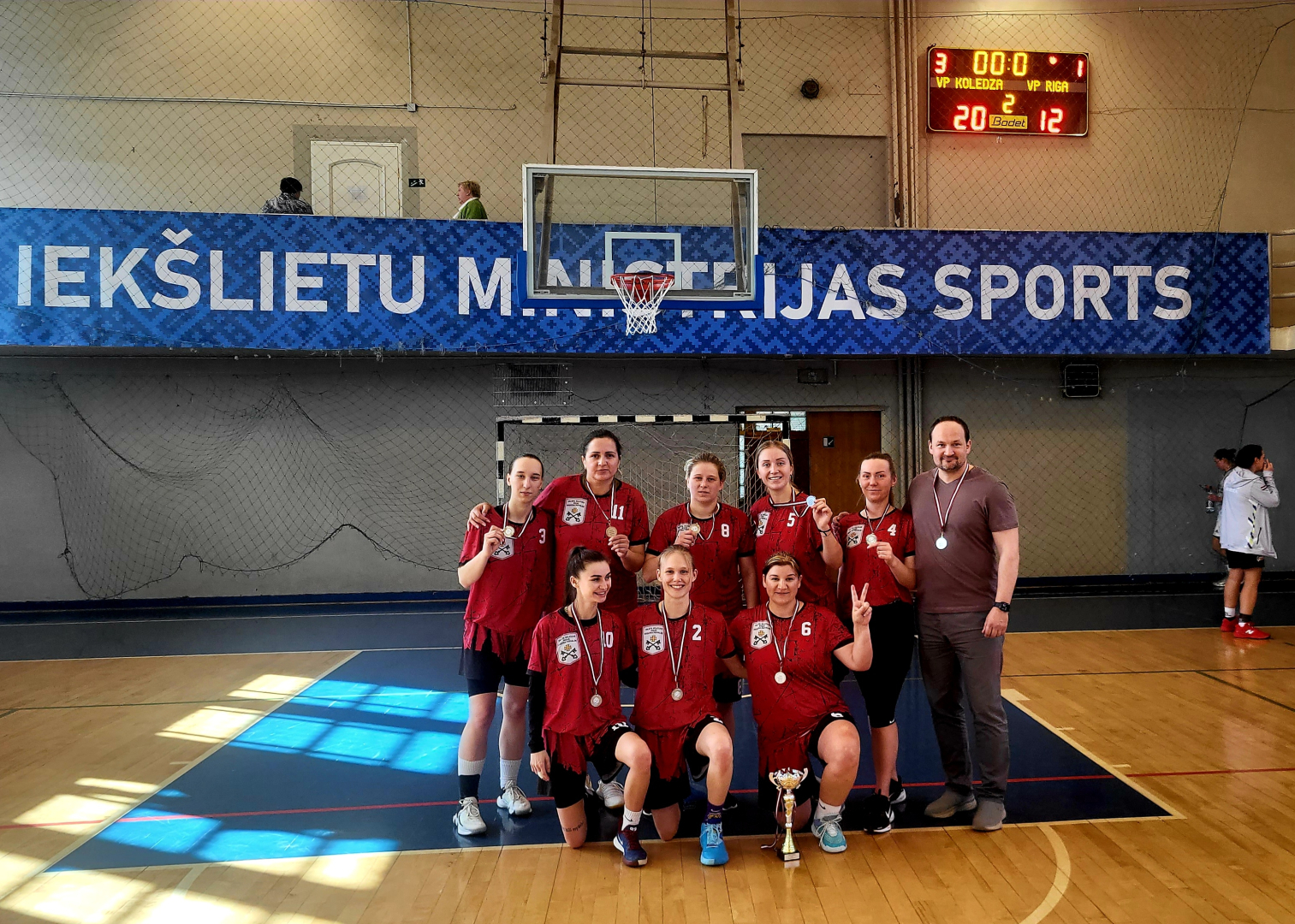 Basketbola sacensību otrās vietas ieguvējas – Valsts policijas Rīgas reģiona pārvaldes komanda ar treneri sporta spēļu zālē. Visām spēlētājām kaklā sudraba medaļas, priekšā uz grīdas – komandas izcīnītais kauss.