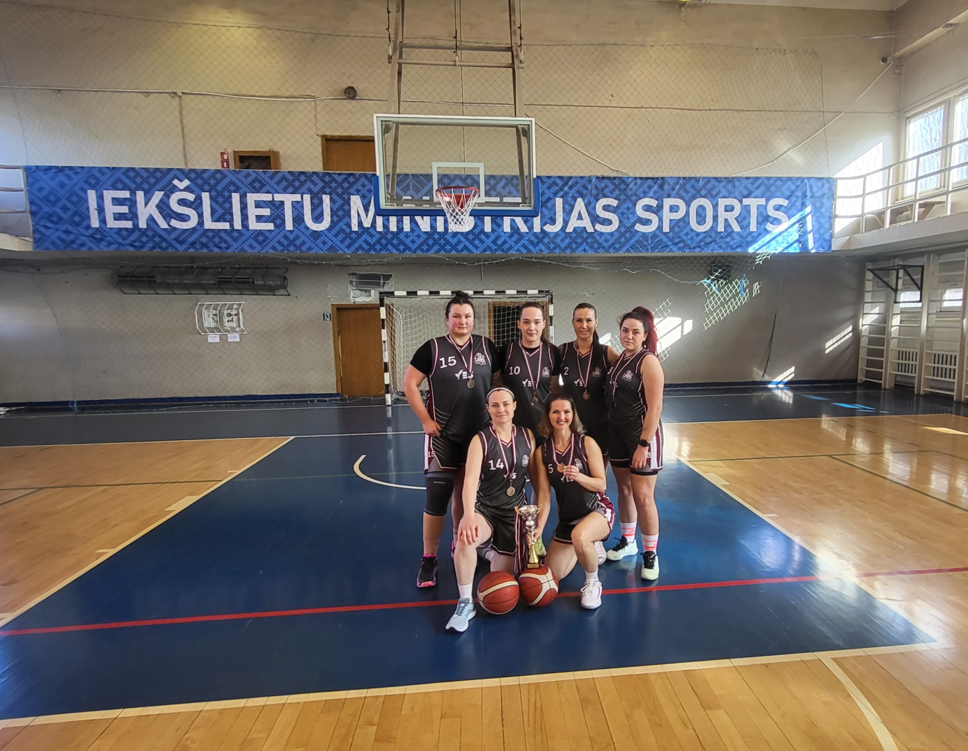 Basketbola sacensību trešās vietas ieguvējas – Valsts robežsardzes komanda sporta spēļu zālē. Visām spēlētājām kaklā bronzas medaļas, priekšā uz grīdas – komandas izcīnītais kauss un divas basketbola bumbas.