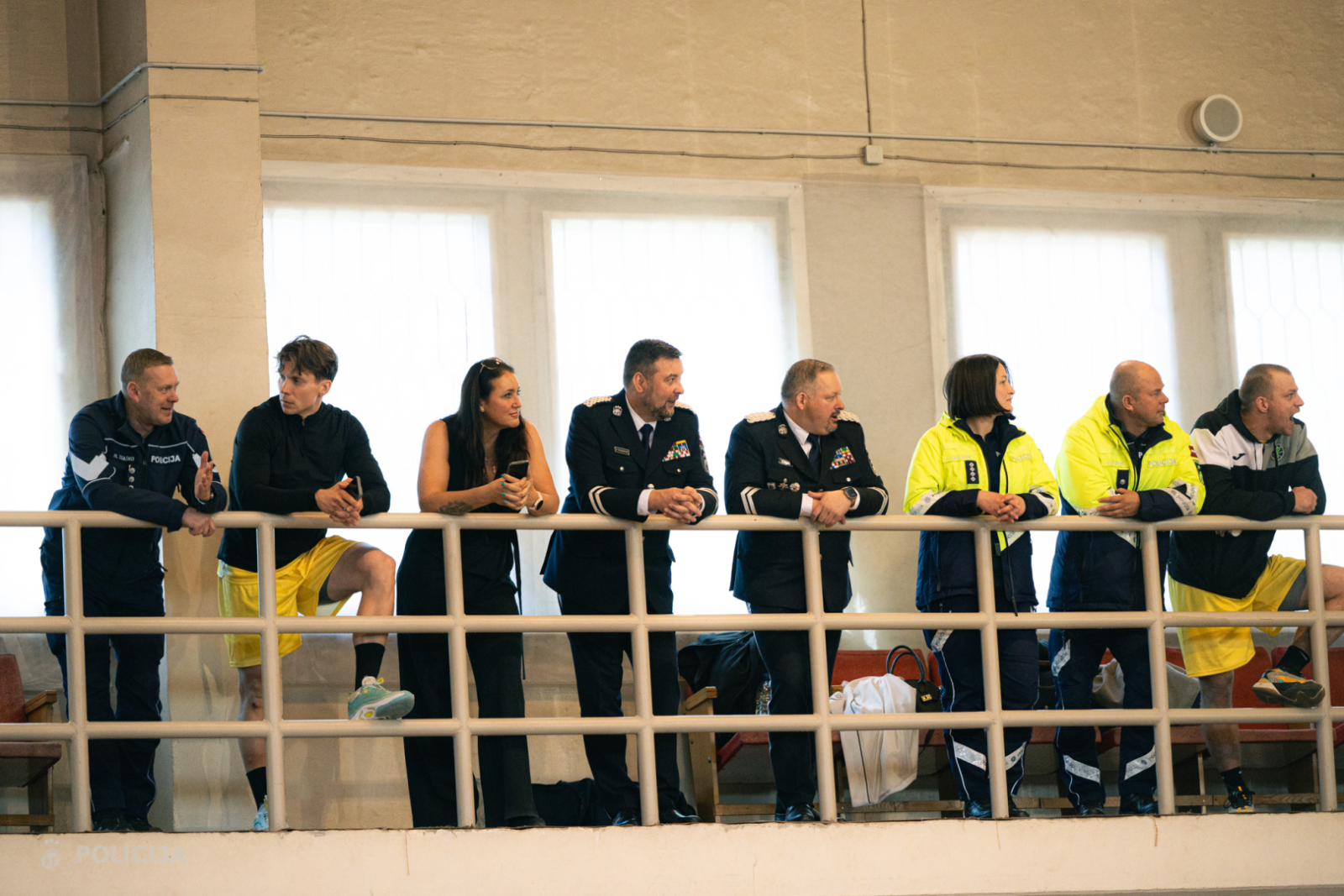 Valsts policijas vadība stāv uz balkona un vēro notiekošo spēli laukumā. 