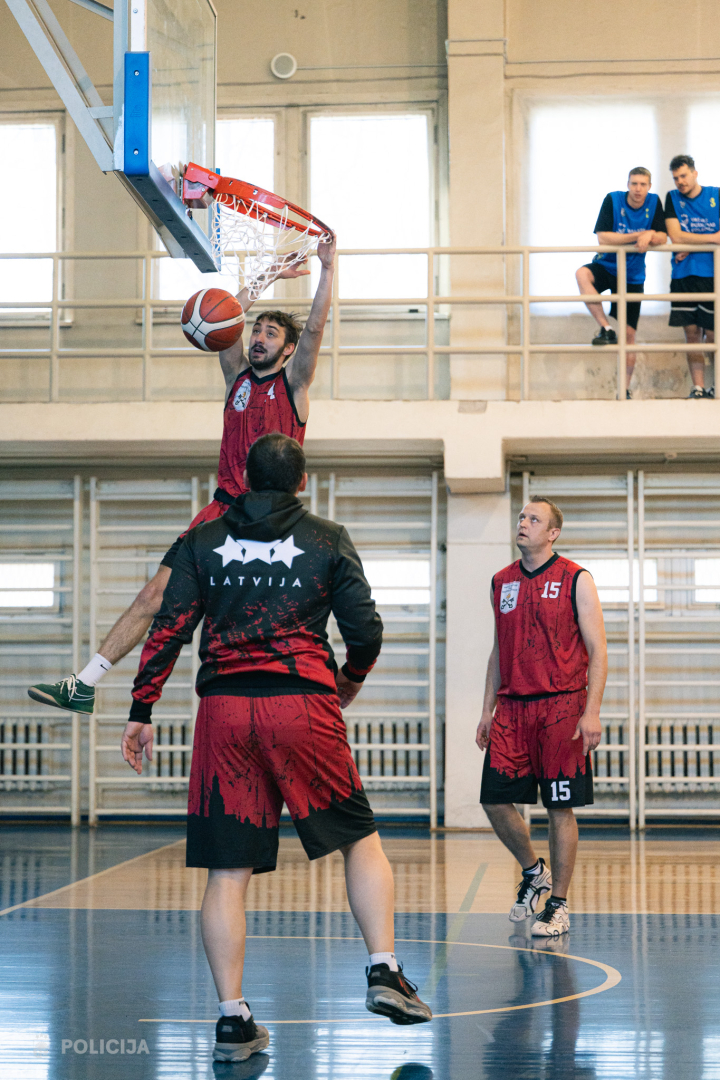 Komandas spēlētājs “izpilda danku”, iemetot bumbu grozā no augšas. Citi spēlētāji un skatītāji vēro epizodi sporta zālē.