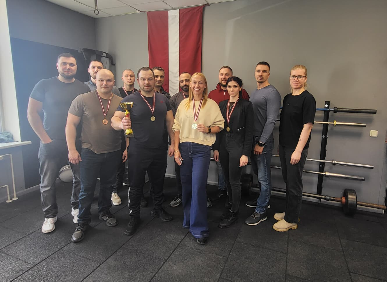 Valsts policijas Kurzemes pārvaldes komanda, 12 cilvēki, 3 sievietes, divas ar medaļām un 8 vīrieši, vienam no viņiem rokās kauss, fonā redzams Latvijas karogs;