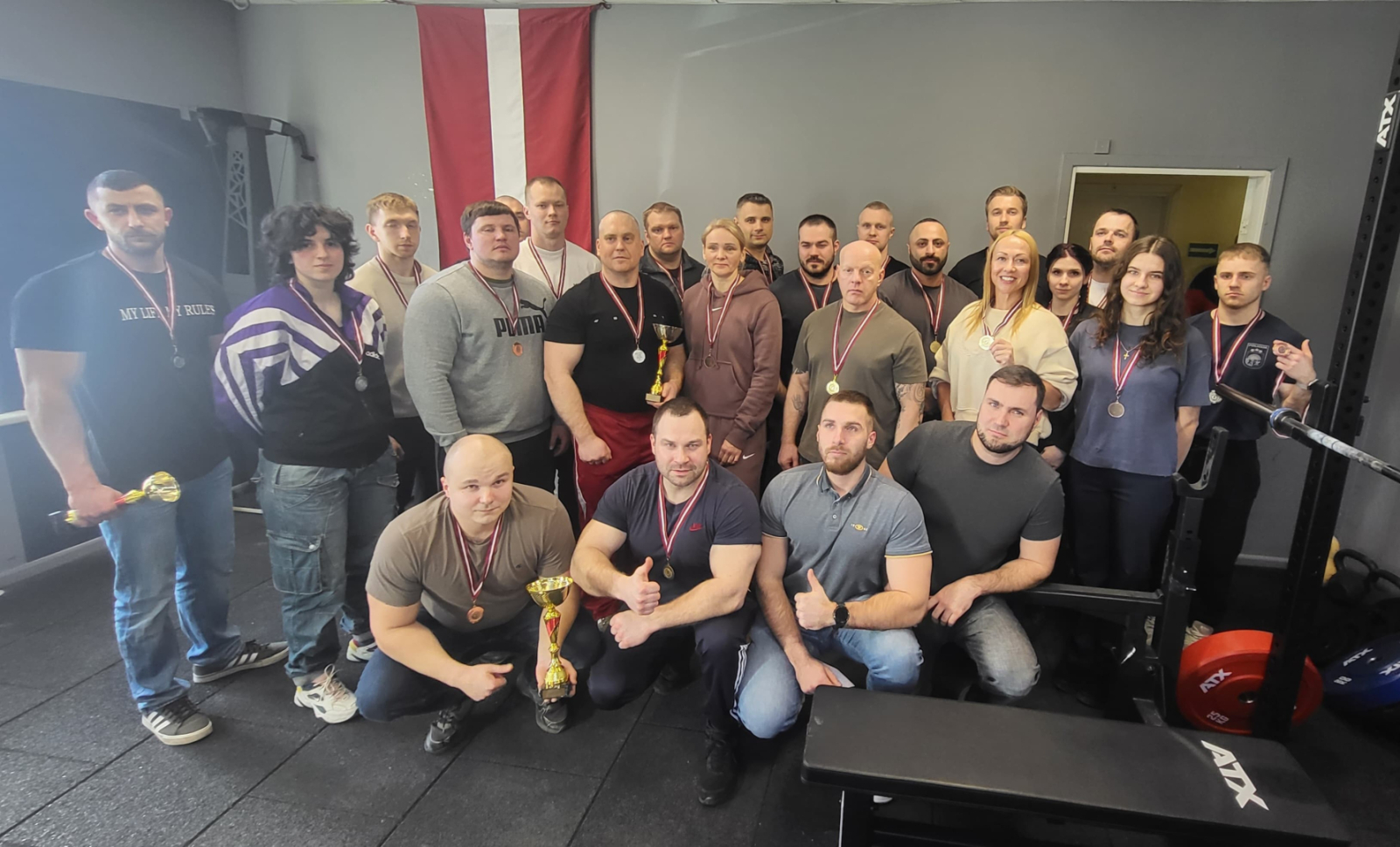 24 cilvēki, 5 sievietes un 19 vīrieši, dažādu komandu pārstāvji, uzvarētāji individuālajā vērtējumā dažādās svara kategorijās, visiem sacensību dalībniekiem kaklā pakārtas medaļas, vienam no vīriešiem otra medaļa ir rokā, trim no vīriešiem rokās ir kausi