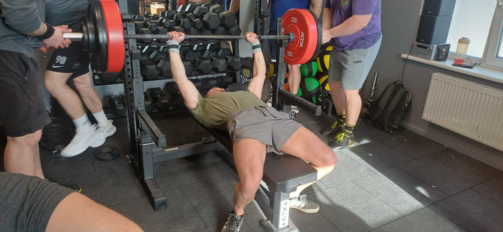 Sacensību dalībnieks spiež svara stieni guļus uz muguras, divi citi dalībnieki drošina, turot stieni aiz galiem