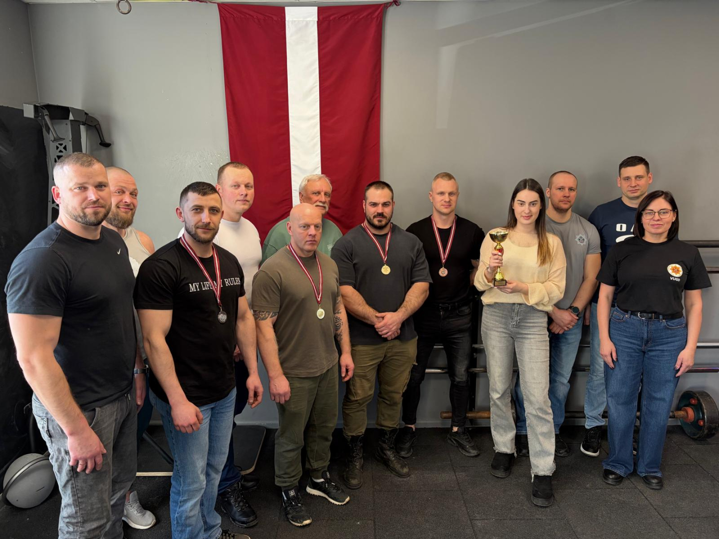 Valsts ugunsdzēsības un glābšanas dienesta komanda, 12 cilvēki, 2 sievietes, 10 vīrieši, viena no sievietēm tur rokās kausu, 4 vīriešiem kaklā medaļas, fonā Latvijas karogs