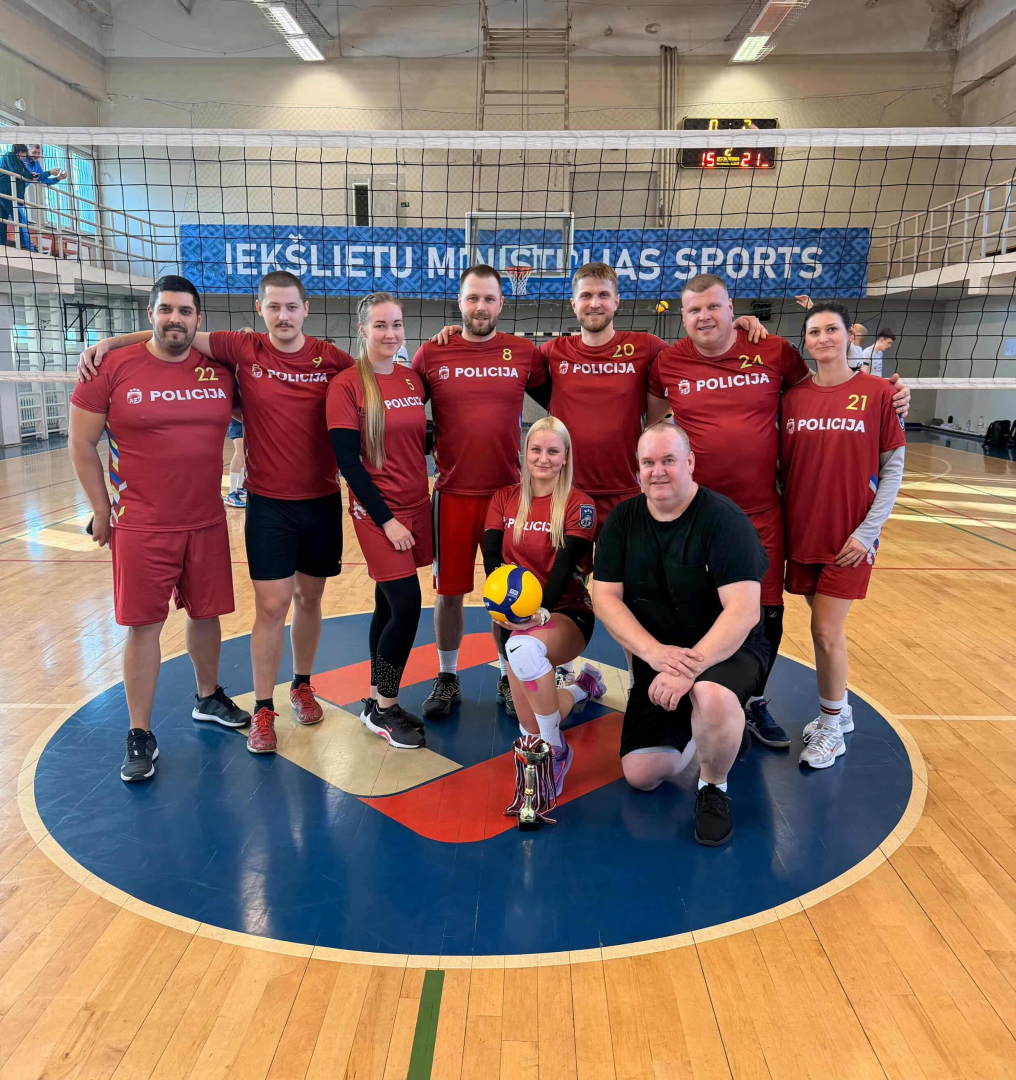 Sporta spēļu zālē volejbola sacensību trešās vietas ieguvēji Valsts policijas Vidzemes reģiona pārvaldes komanda tumši sarkanās formās viens spēlētājs melnā seši vīrieši un trīs sievietes komandai priekšā uz grīdas kauss ar medaļām