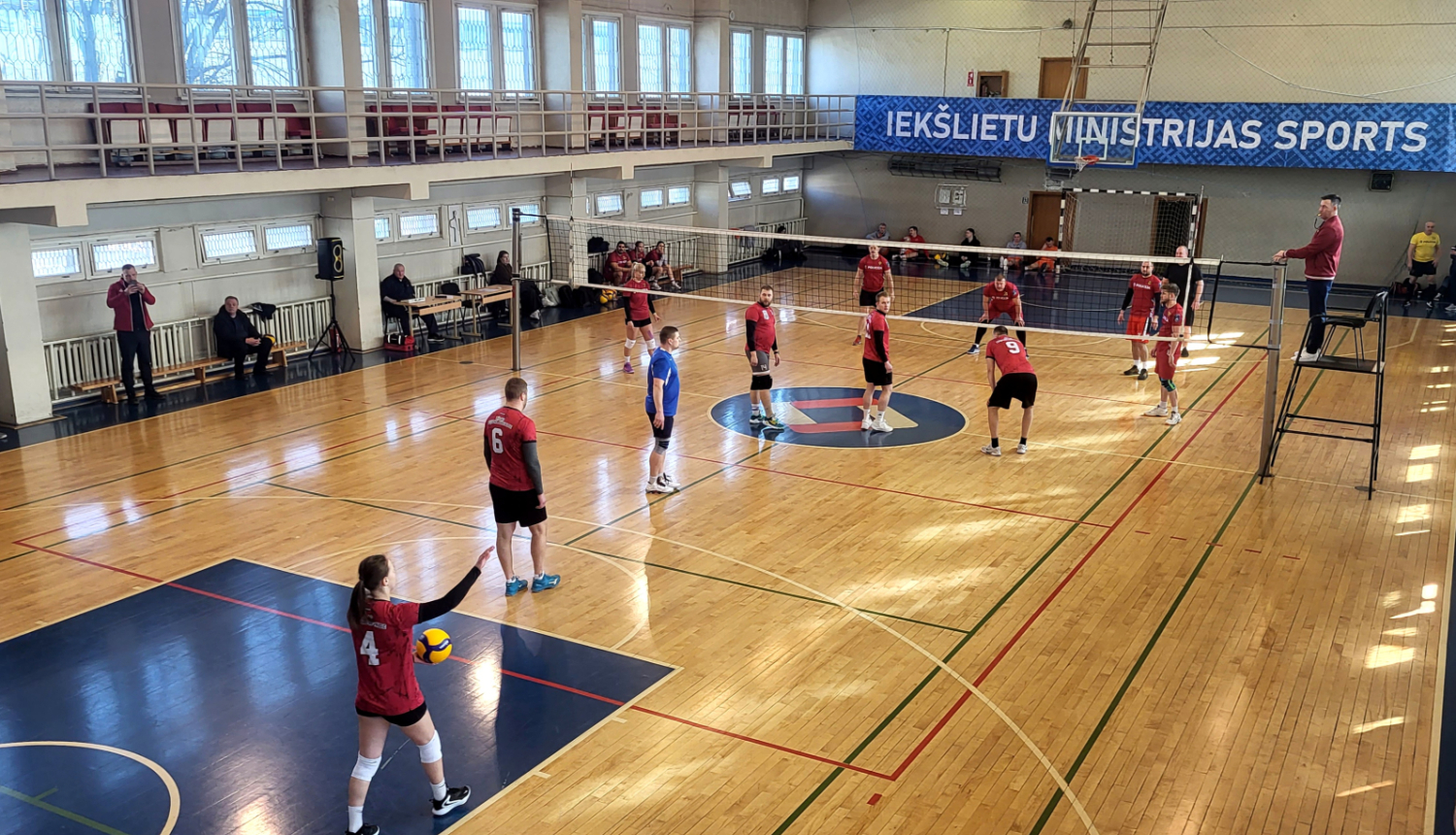 No augšas uzņemta fotogrāfija atklāj intensīvu volejbola maču plašā, saules apgaismotā sporta zālē, kas met asas ēnas uz koka grīdas. Abās volejbola tīkla pusēs pa sešiem spēlētājiem: pieci vīrieši nogaidošās pozās gaida kad sieviete servēs bumbu. Laukuma centrā, uz paaugstinātas platformas tieši pie tīkla, stāv tiesnesis ar svilpi mutē, uzmanīgi seko katram spēles momentam. 