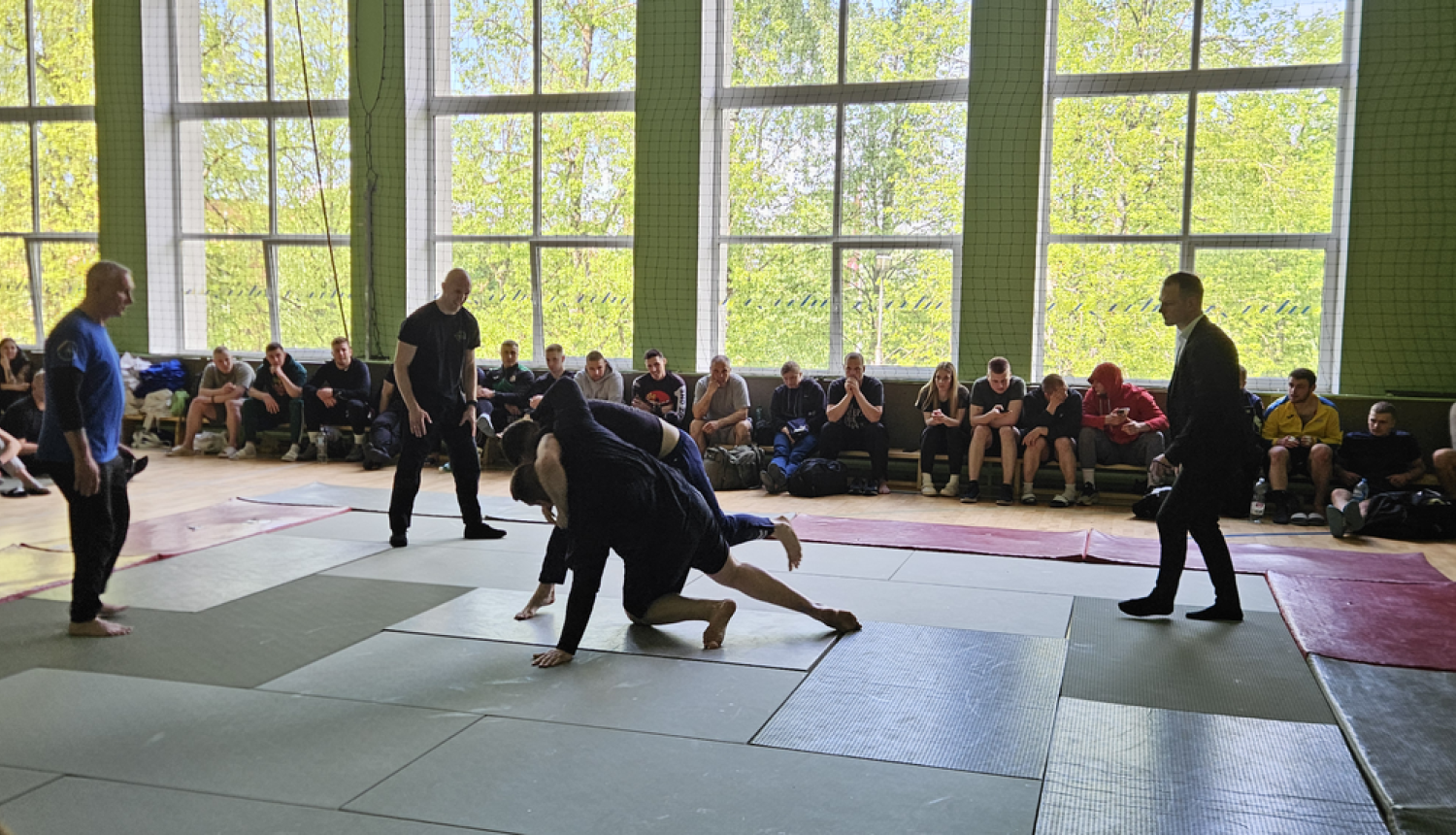 Sporta zālē uz tatami cīnās divi vīrieši, notiek džudo cīņa. Blakus notiekošo vēro tiesnesis un citi sacensību dalībnieki.