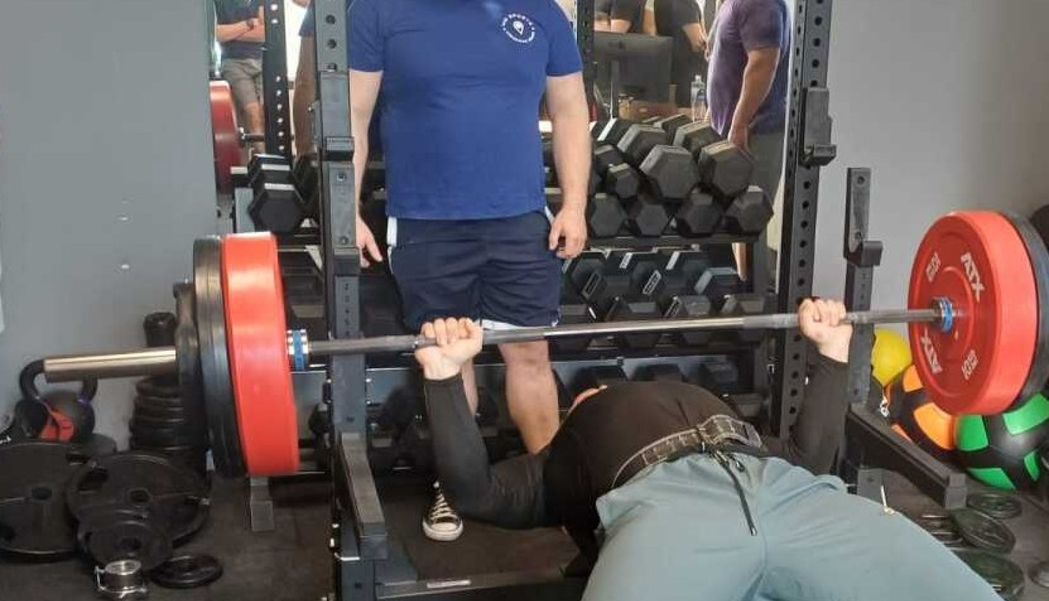 Sporta zālē veic guļus spiešanu ar stieni uz sola.Fonā hanteles.